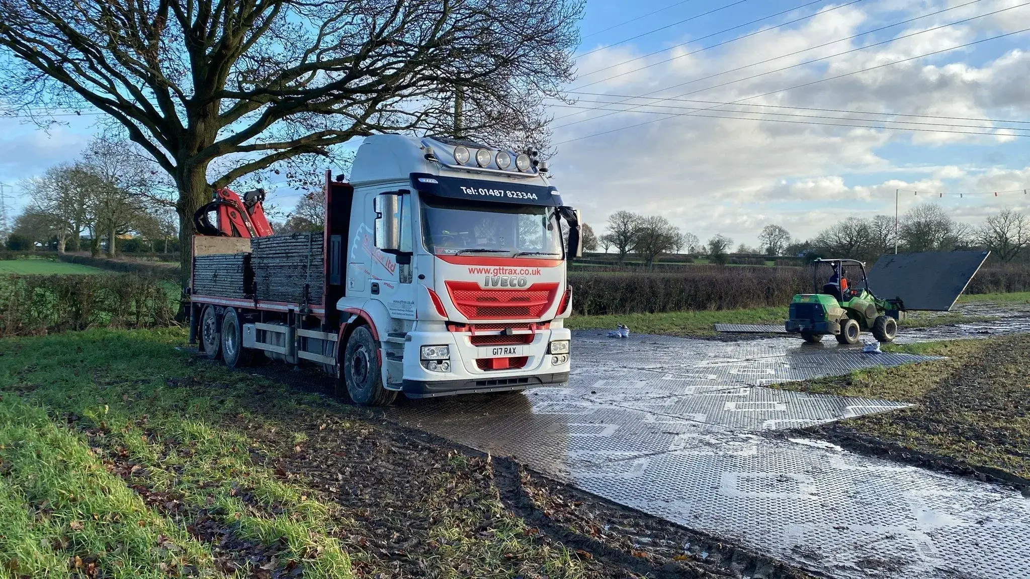 White  truck with crane and green utility vehicle laying temporary PowerTrax 60 mats on a muddy field path.