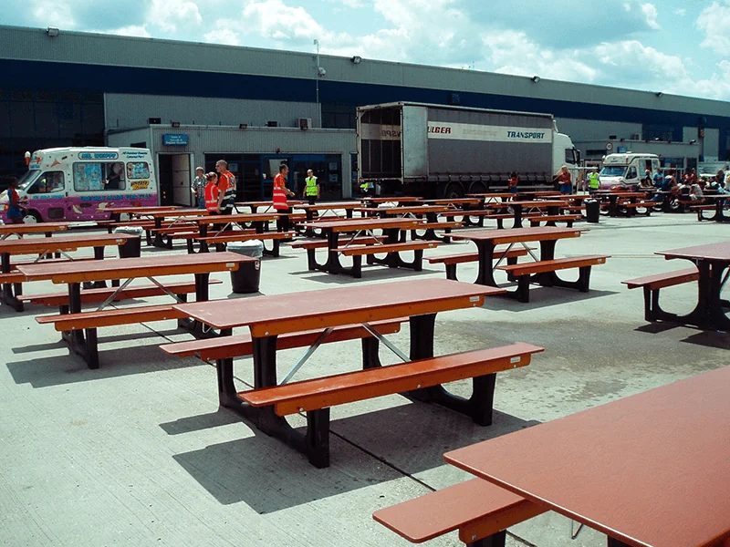 Numerous empty brown FoldTable picnic tables on a paved lot. People, ice cream van, and large truck near an industrial building.