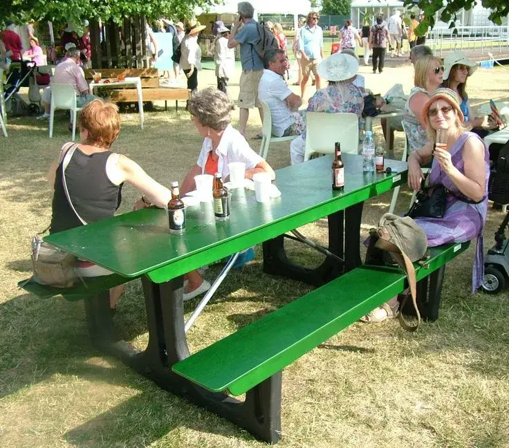 Group of people gathered outdoors around a green FoldTable picnic table, some drinking. One woman in purple smiles at the camera.