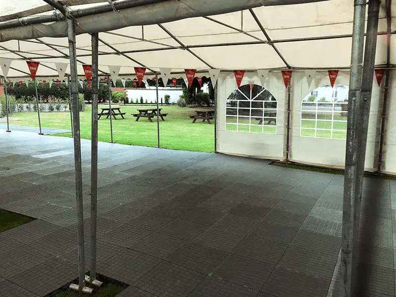 View from inside an empty white event tent, showing a grey tiled LuxTrax floor and red-white banners. A green lawn with picnic tables is outside.