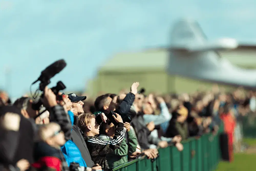 Crowd of people looking up, some with cameras & mic, behind a green fence. Blurry airplane & building in background.
