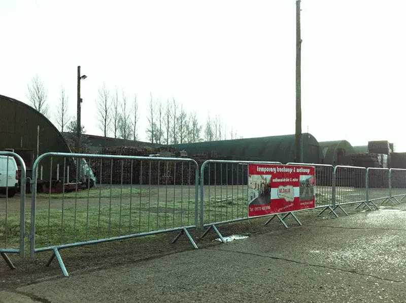 Temporary metal crowd control barriers line an industrial yard with dark, curved buildings, a large log pile, and bare trees.