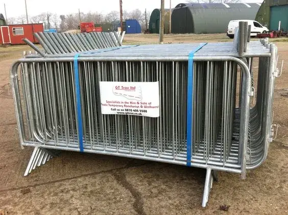 Stack of galvanized metal crowd control barriers, strapped, on dirt ground. Industrial buildings and a white van are in the background.