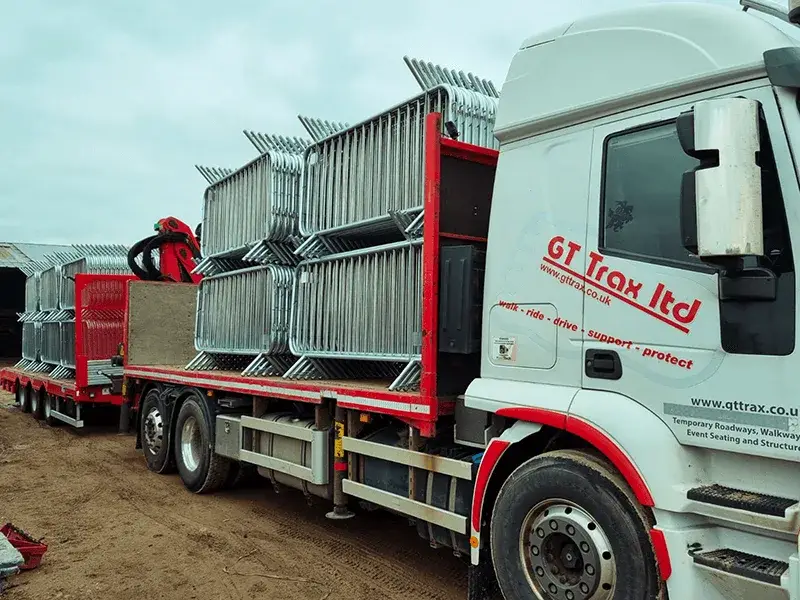 White flatbed truck and trailer loaded with silver metal crowd control barriers on a dirt road.
