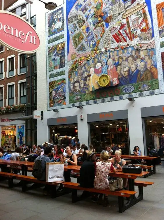 Group of people sitting at FoldTable picnic tables in an outdoor area with a large, colorful mosaic mural and shop facades.