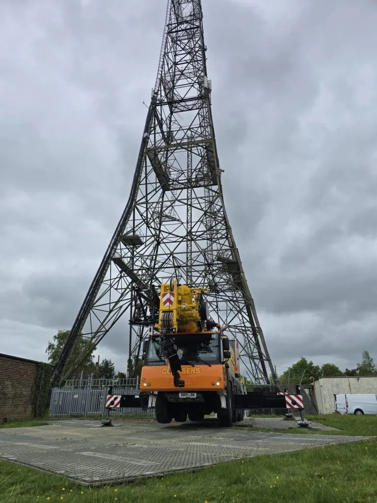Orange mobile crane with outriggers extended on PowerTrax 60 ground mats, positioned at base of tall lattice tower. Cloudy sky.