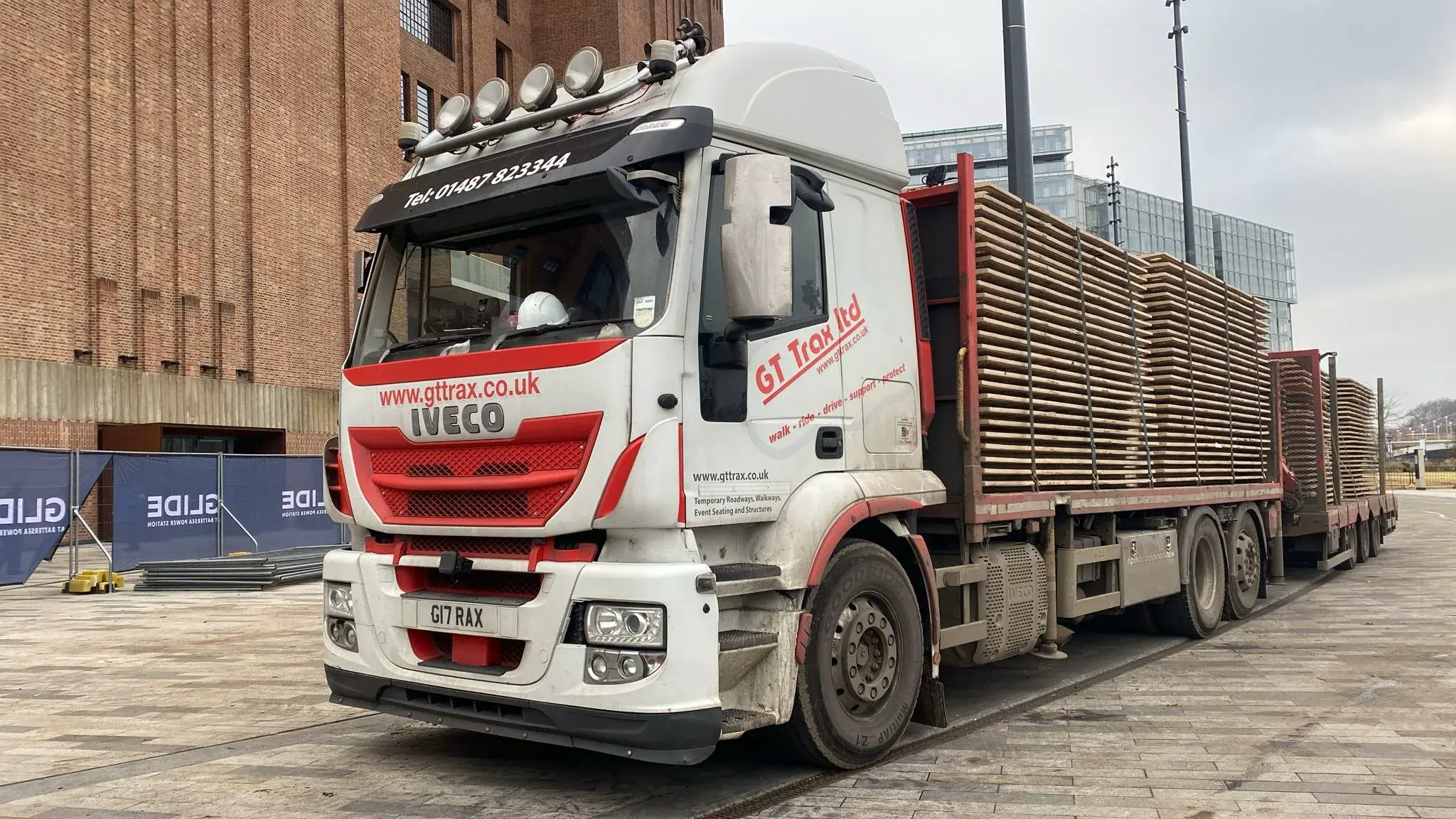 White and red semi-truck with a long flatbed trailer loaded with stacks of PowerTrax 60 temporary ground mats, parked on pavement near a brick building.
