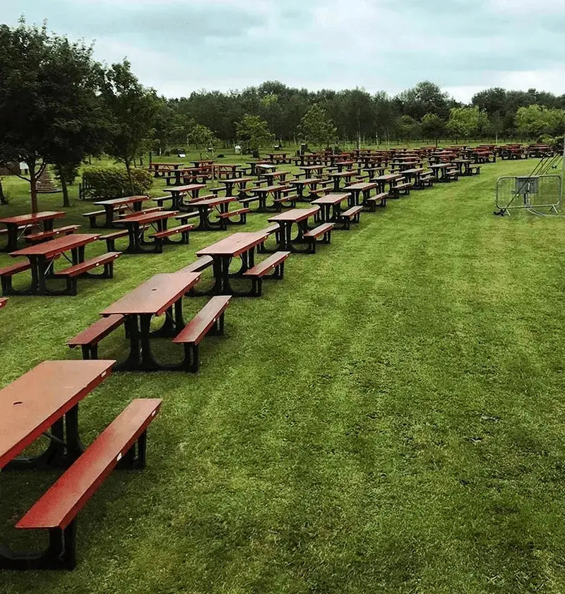 Hundreds of empty red and black FoldTable picnic tables arranged in rows on a large green grass field under a cloudy sky.