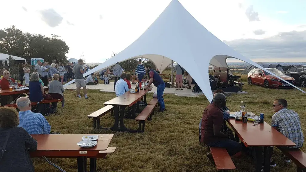 People gather under a white StarShade tent and around FoldTable picnic tables in a grassy field. Cars are visible in the background.