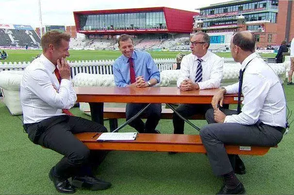 Four men in shirts and ties sit at a FoldTable picnic table on a cricket ground, with stadium stands and a red building in the background.