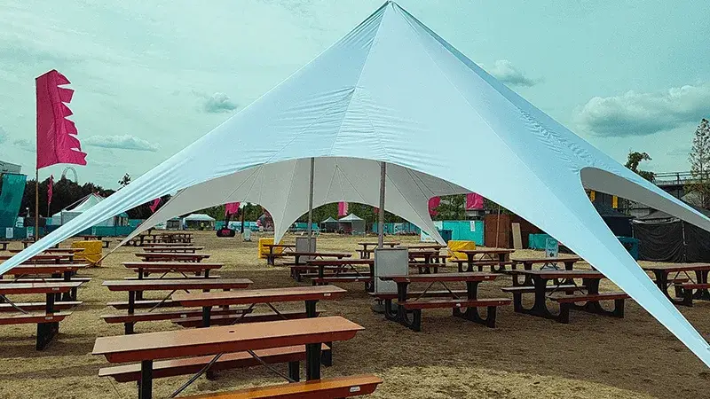White StarShade canopy covers rows of empty wooden FoldTable picnic tables on a dry outdoor field under a cloudy sky.