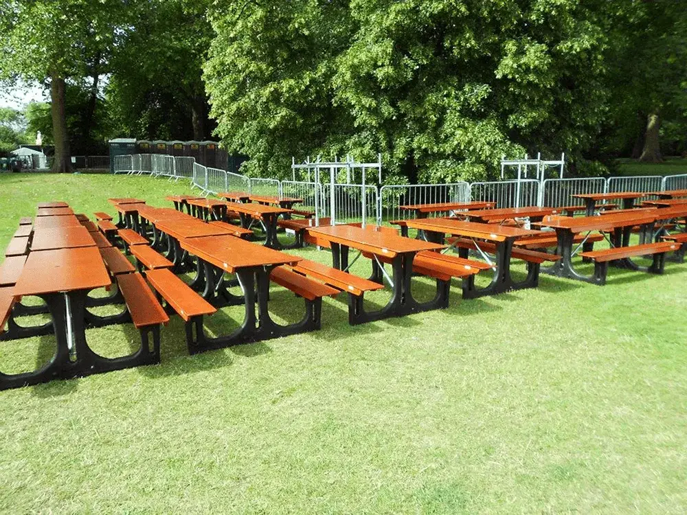 Rows of brown FoldTable picnic tables with black bases on a grassy field. Trees, metal crowd control barriers, and portable toilets are in the background.