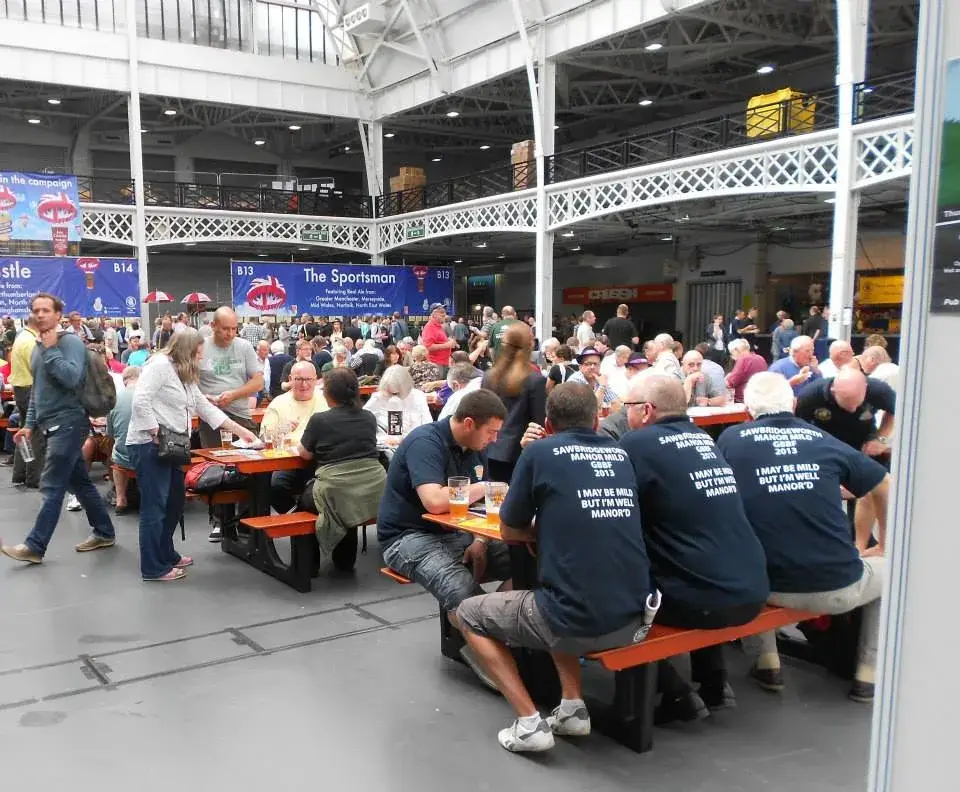 Crowd of people socialize and drink beer at orange FoldTable picnic tables in a vast indoor hall. Some men wear matching shirts.