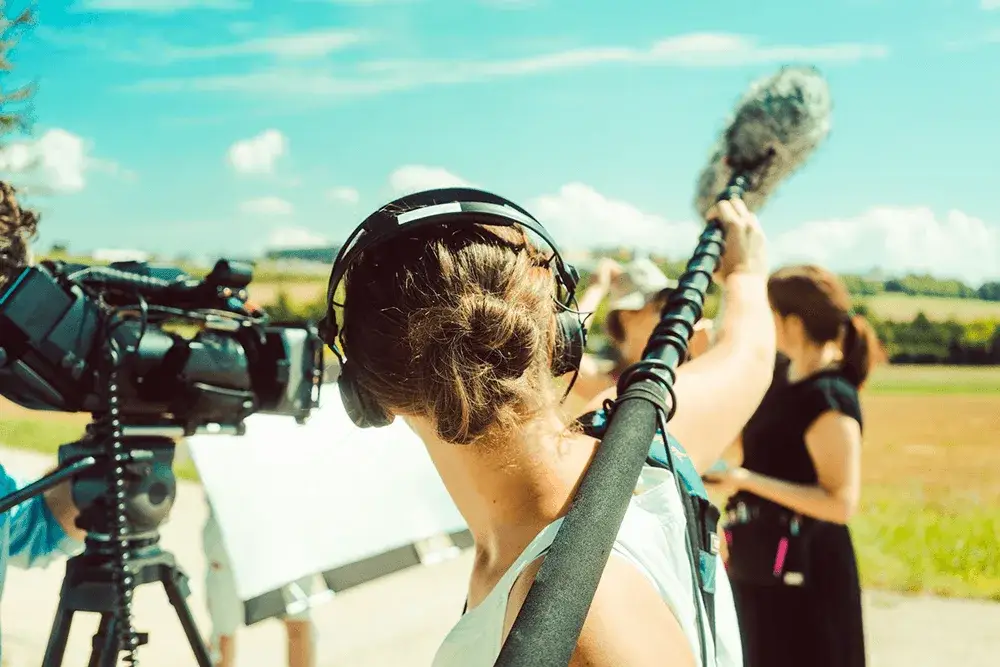 Boom operator with headphones holds a mic on a film set outdoors. Video camera on tripod, blue sky, and fields in background.