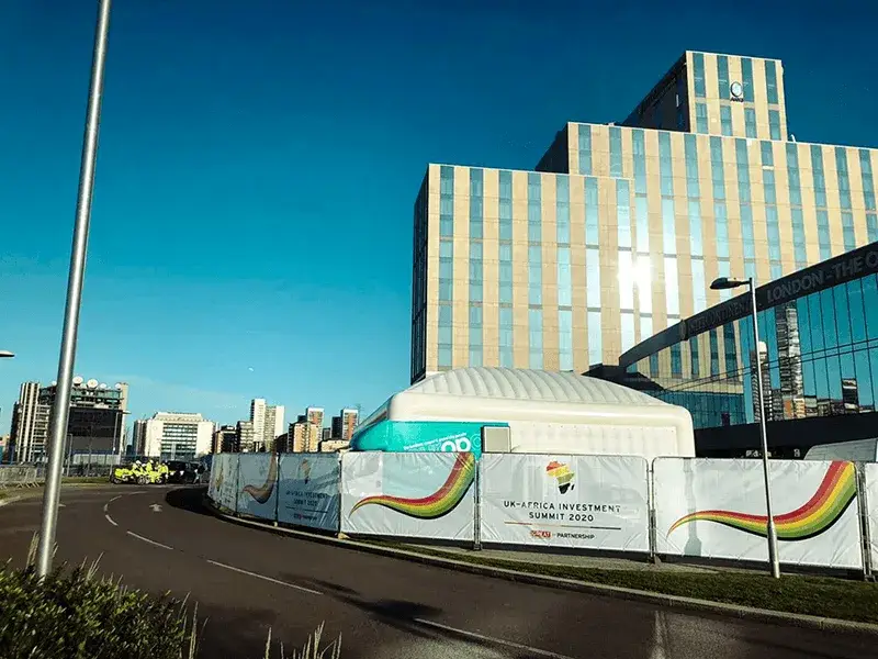 Banners on Heras fencing in front of white tent, and modern building under clear blue sky.