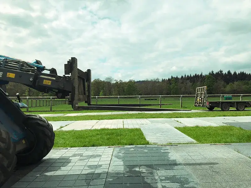 A telehandler with forks extended over grass on PowerTrax 20 temporary flooring, a flatbed trailer in the distance. Trees and cloudy sky, paved foreground.