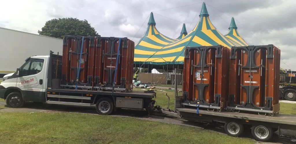 White truck and trailer loaded with large brown folded FoldTable picnic table on grass, with a yellow and green striped circus tent behind.