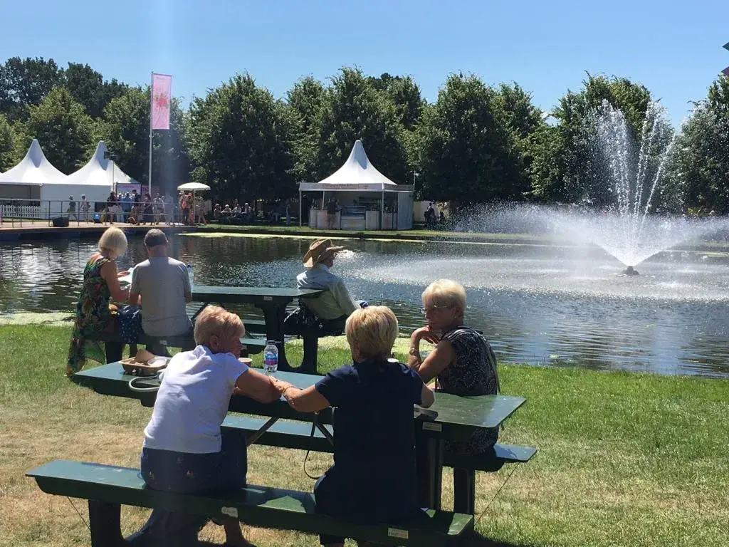 Older adults sit at FoldTable picnic tables beside a pond with a fountain, white tents, and trees on a sunny day.