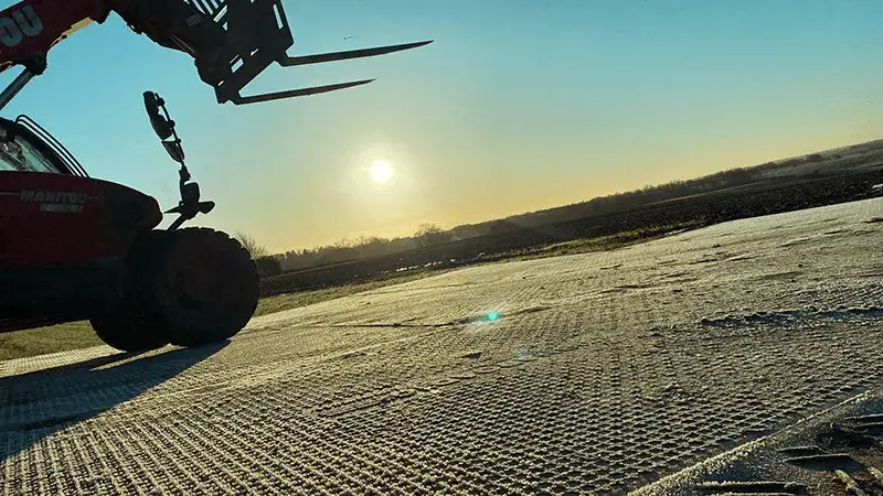 Red Manitou telehandler with forks up on textured PowerTrax 60 ground mats, sun low in sky over distant fields.