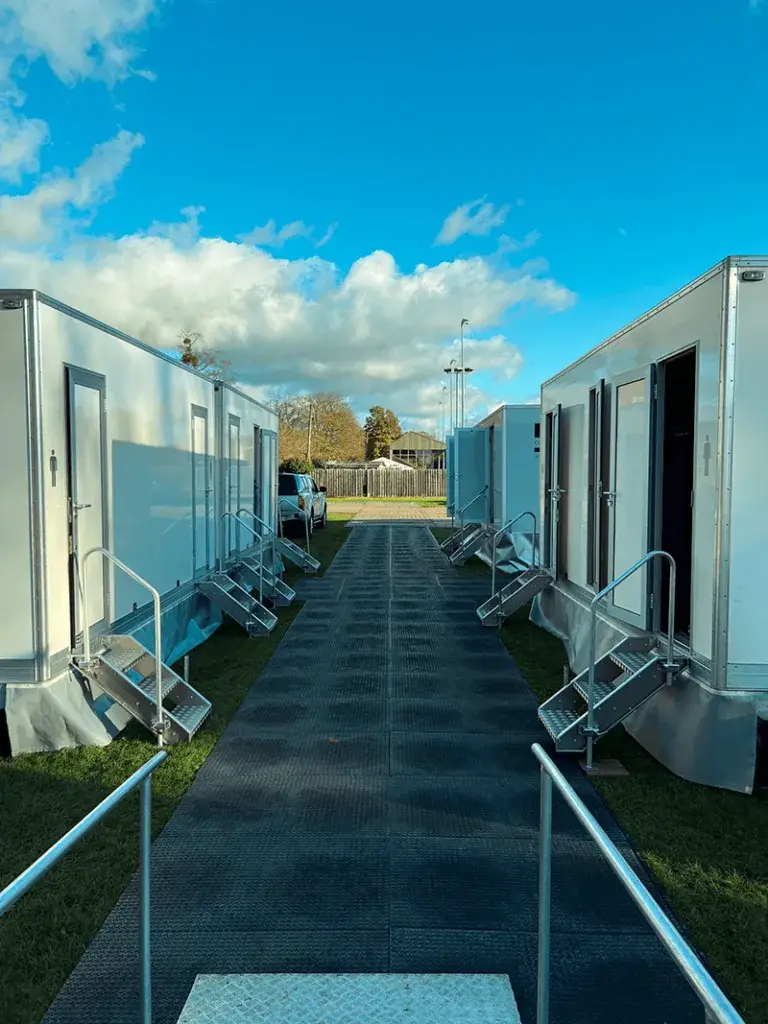 A path of dark WalkOver mats between two lines of white portable toilets with stairs and handrails, under a blue sky with white clouds.