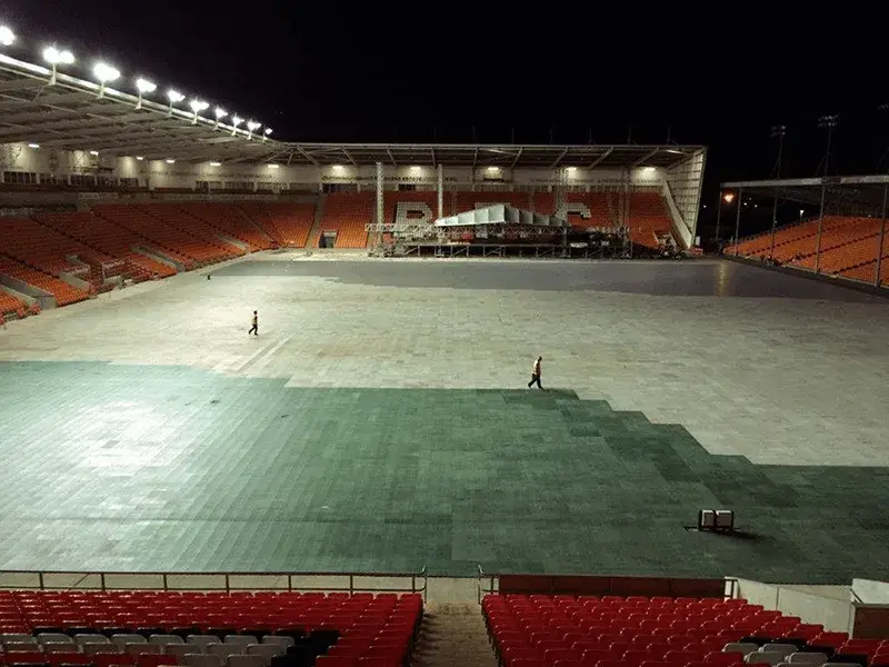 Empty stadium at night with two people walking on a field partly covered with green LiteTrax ground mats. Orange and red seats visible.