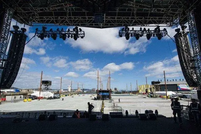 View from concert stage: hanging speakers, lights, crew on empty concrete outdoor venue under blue sky with clouds.