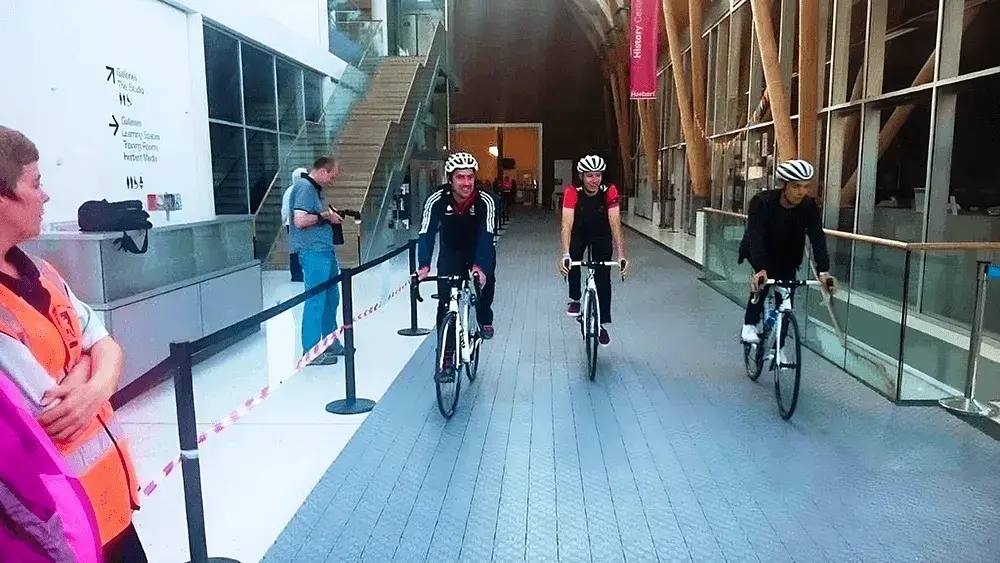 Three cyclists wearing helmets ride road bikes down a path of LiteTrax pathway inside a wide indoor hallway. A woman in a safety vest watches them.