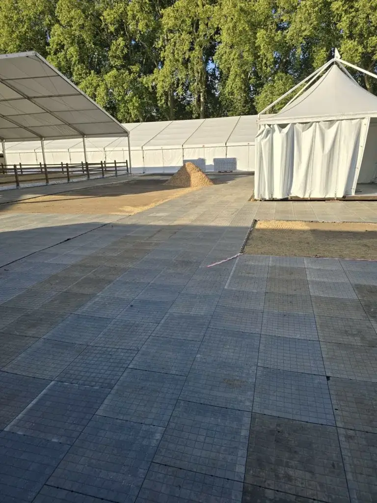Large outdoor event space with white tents, dark grey LuxTrax interlocking floor tiles, dirt, a pile of gravel, and green trees.