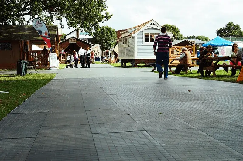 Man walks on a tiled LuxTrax path through an outdoor fair with stalls, tiny houses, and people sitting at picnic tables under trees.