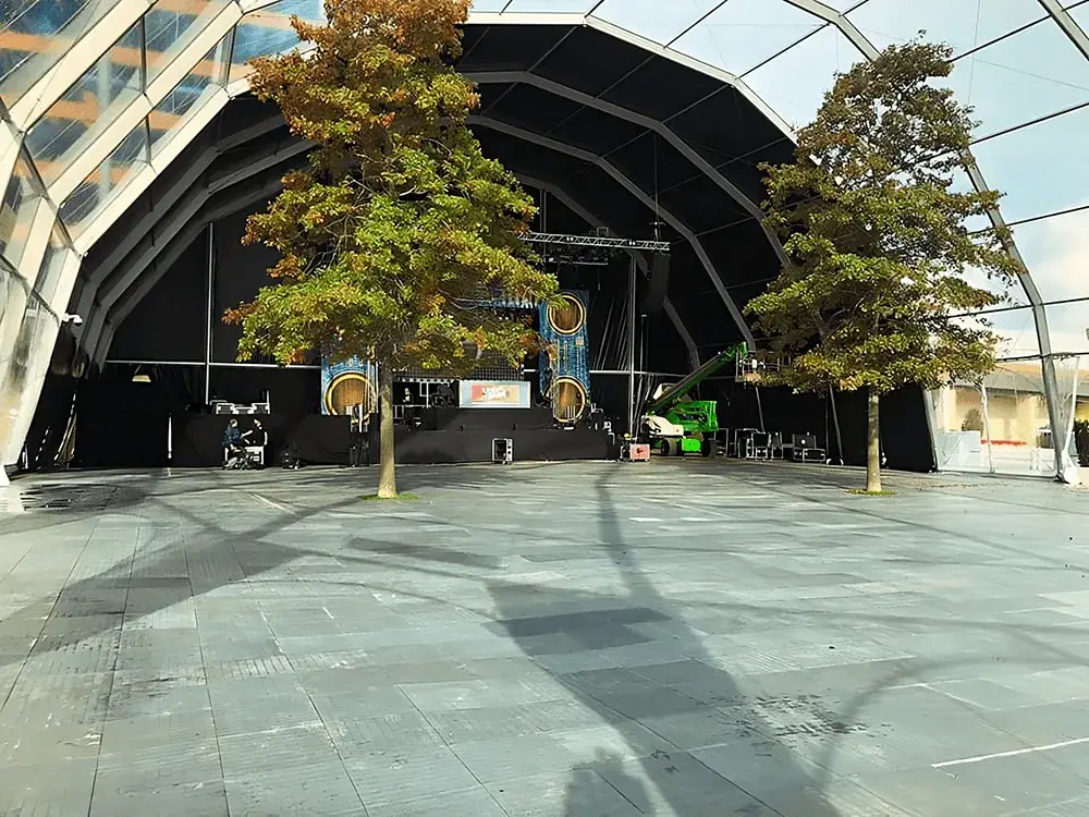 Large arched event tent interior with two trees on a paved LuxTrax floor. Stage setup, a green lift, and a worker are at the back.