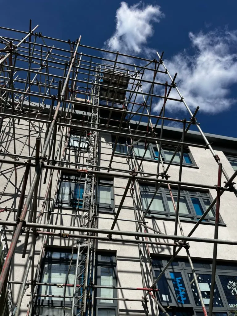 Scaffolding covers the front of a multi-story building with windows, set against a blue sky with white clouds.