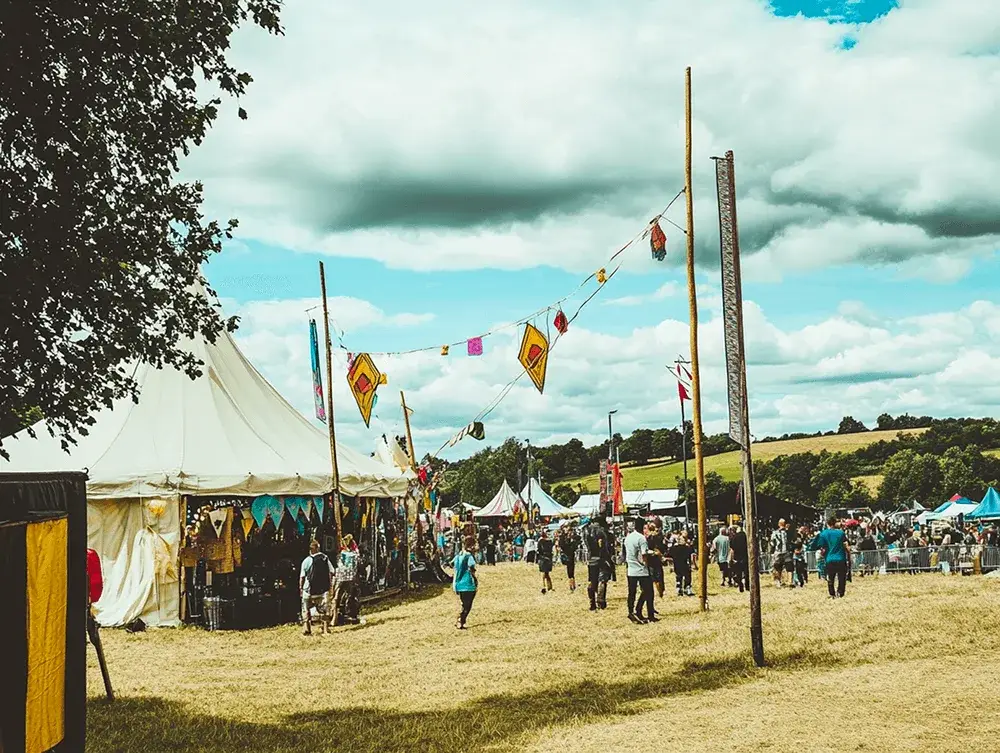 Grassy field outdoor festival with white vendor tents, a crowd of people, colorful bunting, and distant hills under a cloudy sky.