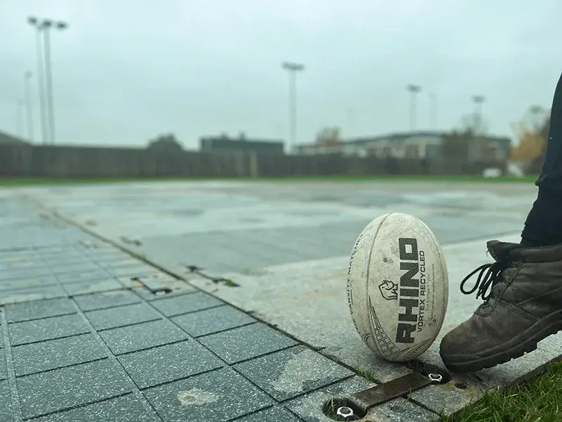 A person's foot in a brown boot next to a dirty white Rhino rugby ball on a wet, grid-patterned PowerTrax 20 trackway surface. Overcast field.