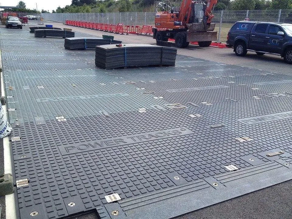 Grey textured PowerTrax 40 ground mats, some labeled "POWER TRACKS", cover the ground. Stacks of mats, an excavator, and a truck are visible.