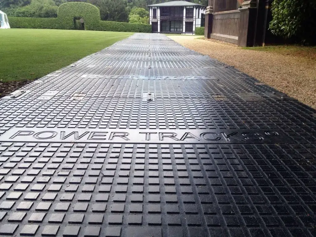 Black textured PowerTrax 40 mats forming a pathway across a grassy lawn towards a modern building. "POWER TRACKS" is visible on the mats.