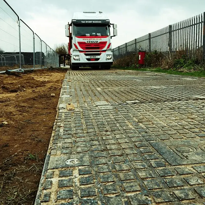 White & red truck on a PowerTrax 40  temporary road of dark gray interlocking mats. Dirt, metal fences, and a red post in the background.