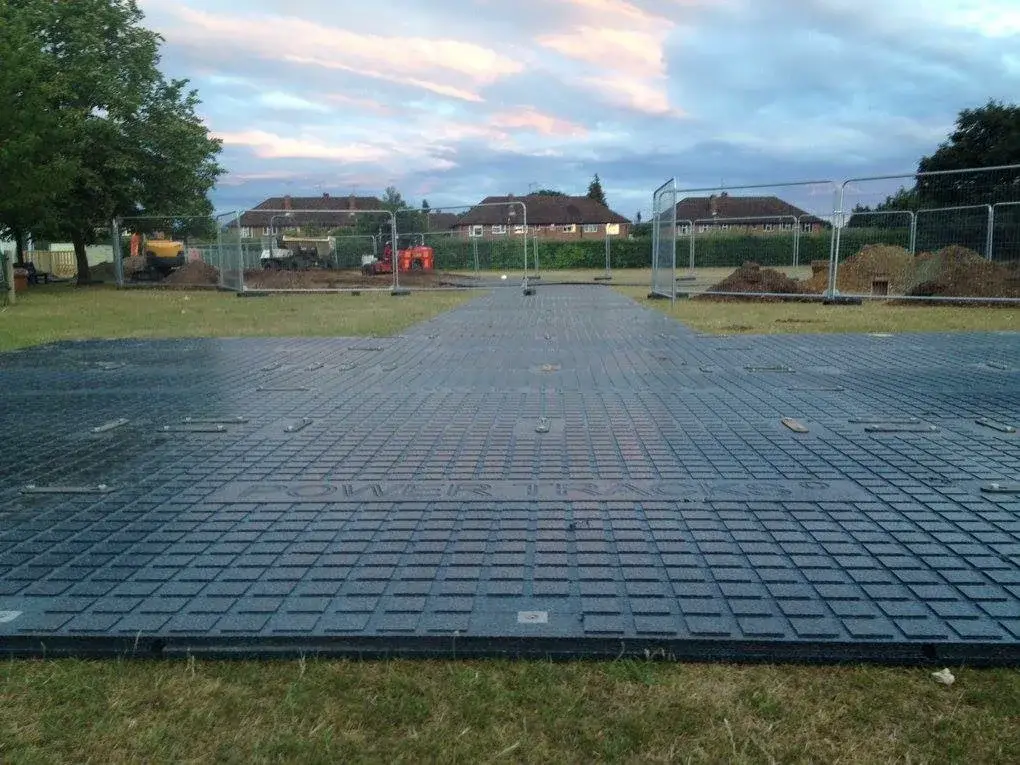 T-shaped path of black PowerTrax 40 temporary ground mats on grass. Construction site with diggers and dirt piles fenced off in background under cloudy sky.