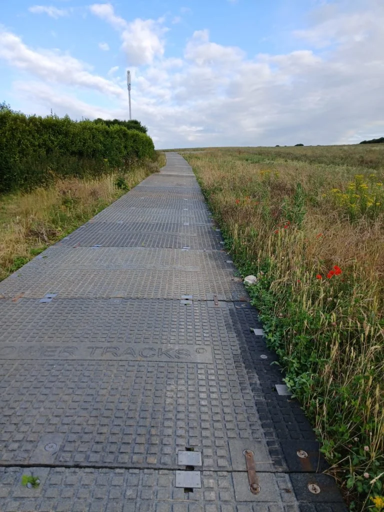 Textured grey PowerTrax 40 track, marked "POWER TRACKS", leads uphill through wild grass and a hedge. A white mast is visible under a blue, cloudy sky.