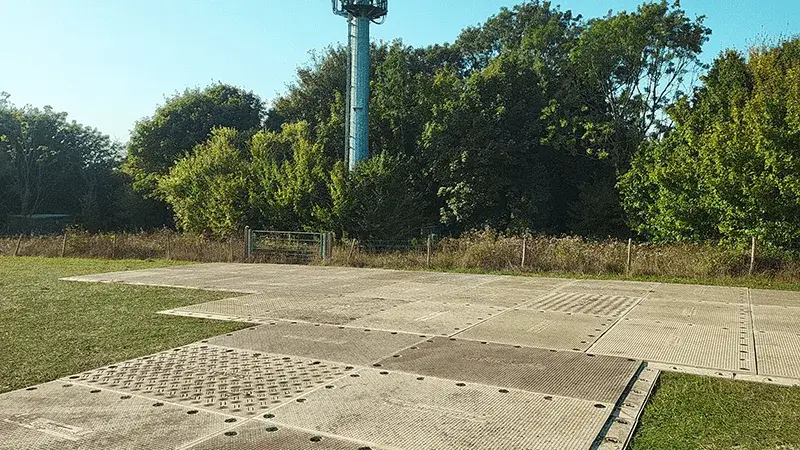 Interlocking grey PowerTrax 60 mats cover a grassy area. A tall tower is visible over dense trees and a fence in the background.