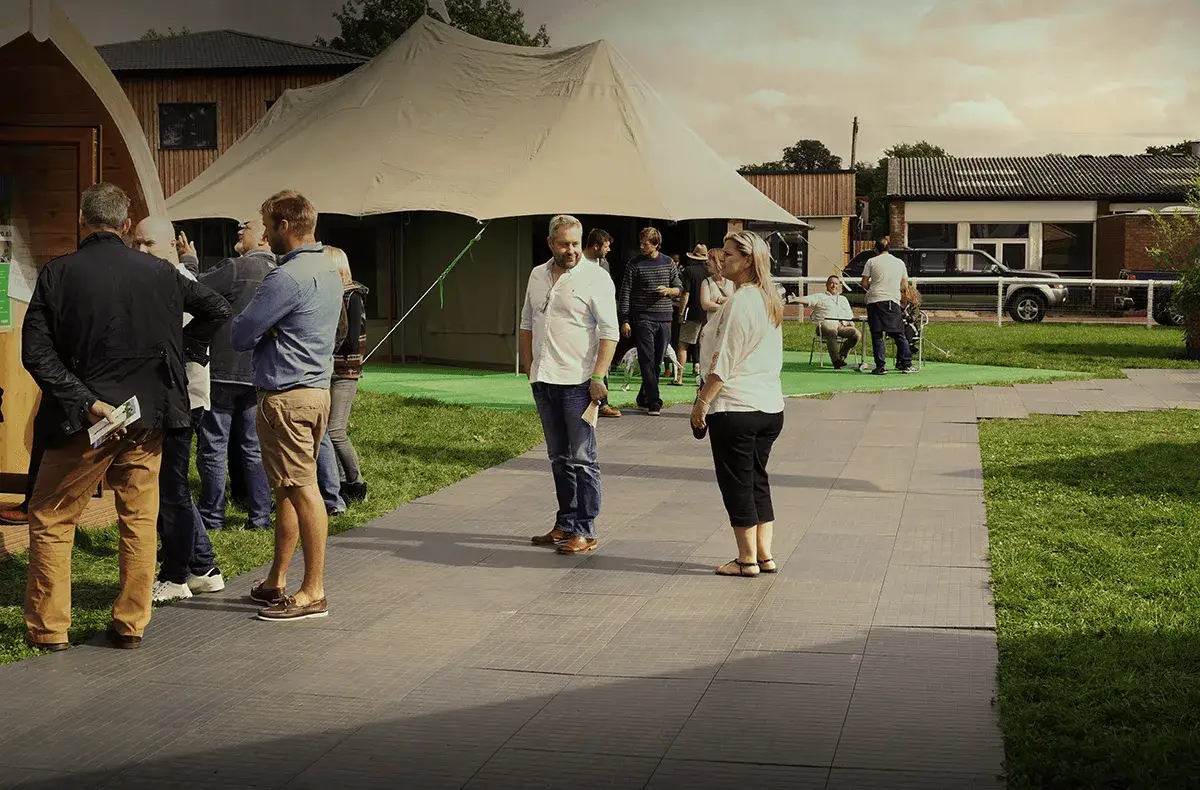 People stand and talk on a tiled LiteTrax path next to a large tent and wooden building. Grass, other buildings, and a parked truck are visible.