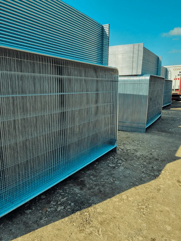 Stacks of new galvanized wire Heras metal temporary fencing panels on a sunny dirt lot under a blue sky.