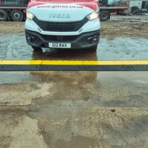 Front of a white van stopped before a black row of cable protections with yellow top on wet concrete ground.