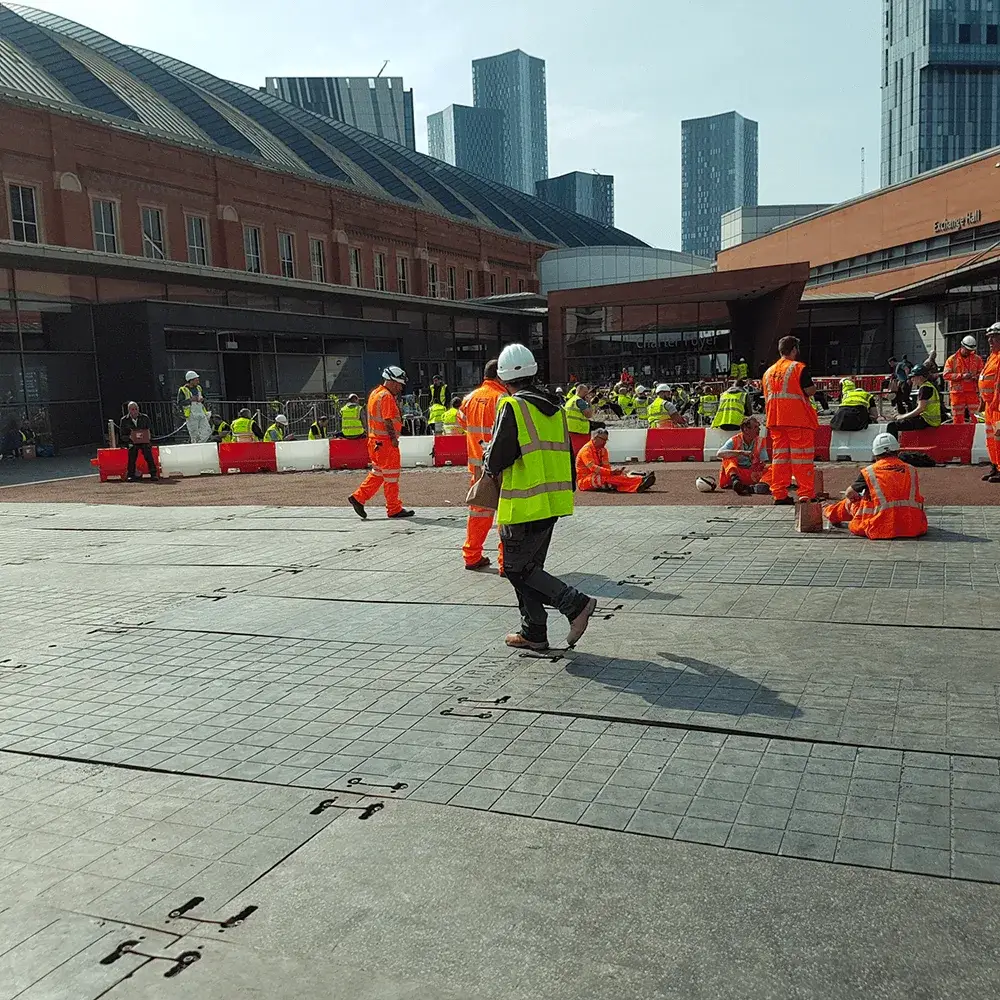 Many construction workers in hi-vis orange and yellow gear on temporary PowerTrax 20 ground mats outdoors. Some walk, many rest. City buildings.