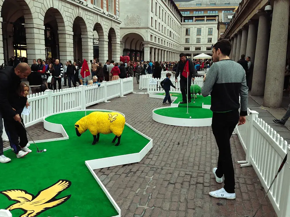 People play mini golf with sheep statues on an outdoor cobblestone square, enclosed by picket fencing, surrounded by historic buildings.