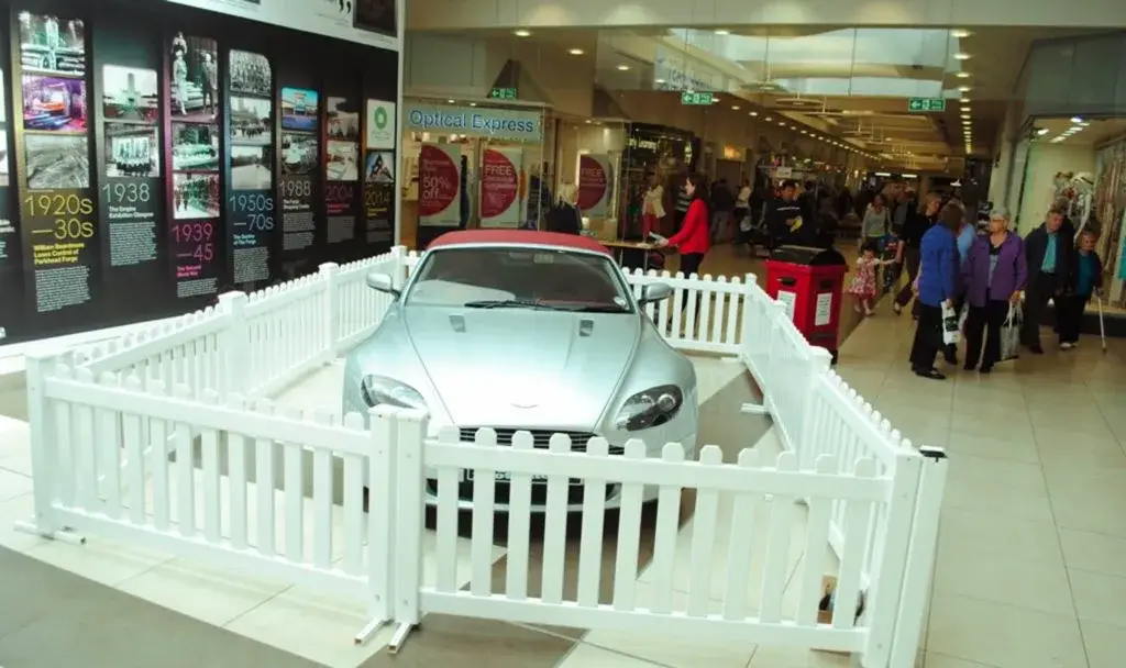 Silver convertible car with red soft top on display behind white picket fence in a busy mall, with shoppers.