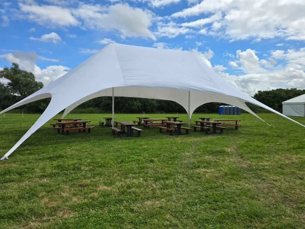 White StarShade canopy tent shading wooden picnic tables in a grassy field under a cloudy sky. Distant trees, porta-potties.