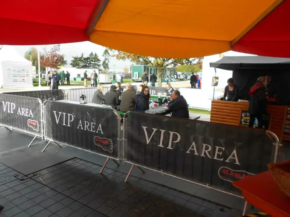 Attendees sit at a table in an outdoor VIP area under a red and orange tent canopy, behind crowd control barriers. Other people stand in the background.