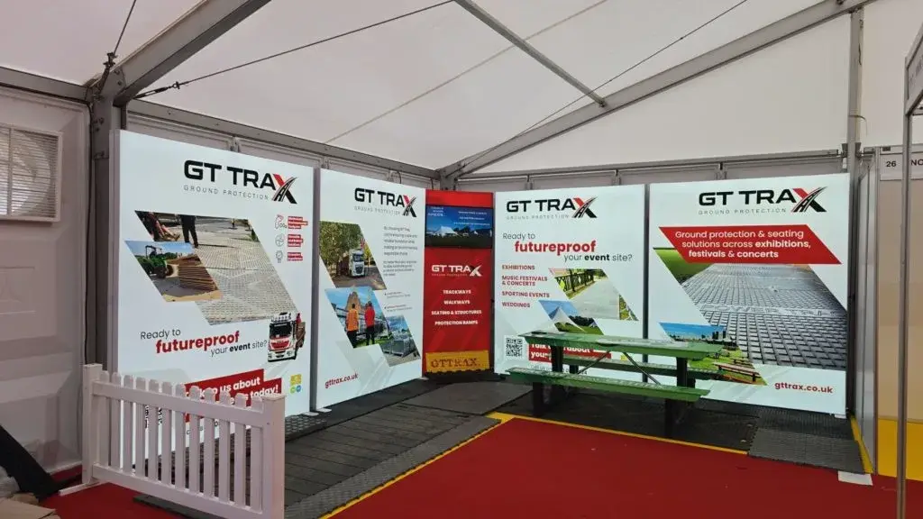 An exhibition stand for GT Trax Ground Protection inside a white event tent, with large banners, red carpet, and a picnic bench.