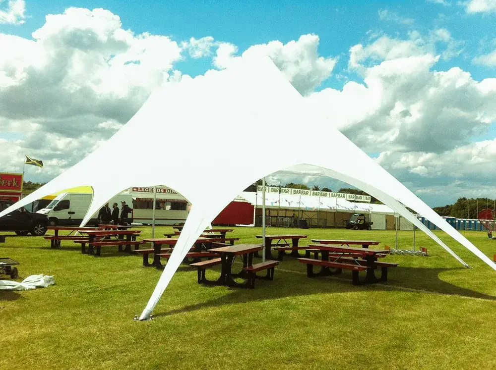 Large white StarShade tent providing shade for FoldTable picnic tables on a grassy field with stalls & tents under a cloudy sky.