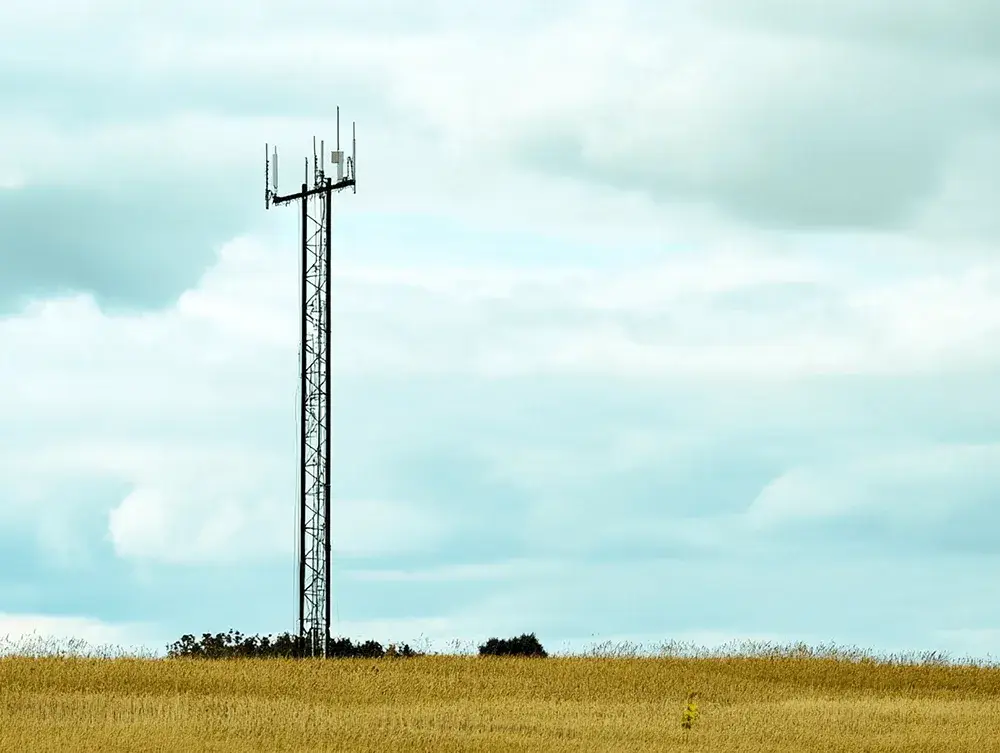 A tall cell tower with antennas stands in a golden field beneath a light blue cloudy sky.